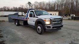 Tow truck parked on a concrete lot with trees and buildings in the background.