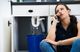 Woman on phone with a leaking sink pipe, water pouring into a bucket under the cabinet.