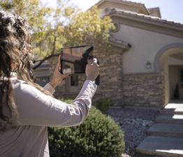 Person using a tablet to take a photo of a house, with trees and sunlight in the background.