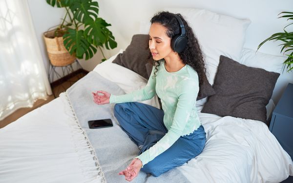 Frau sitzt mit Kopfhörern auf dem Bett und wendet ein Entspannungsverfahren an, um muskulären und seelischen Problemen entgegen zu wirken.