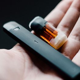 A hand holding a vape pen with an orange cartridge on a dark background.