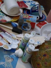 Malutensilien und bemalte Tassen auf einem Tisch, darunter Pinsel, Farben und Tonfiguren.