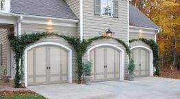 Three garage doors with ivy growing on a house, set against a backdrop of autumn trees.