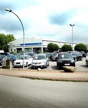 Autohandel mit verschiedenen Fahrzeugen auf dem Parkplatz, umgeben von Bäumen und Wolken.