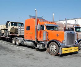 Orange semi-truck transporting military vehicles on a flatbed trailer in a parking lot.