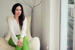 Eine Frau sitzt auf einem Stuhl, trägt eine weiße Bluse und grüne Hose.