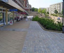 Fußweg mit Geschäften und Cafés links, Bäumen und Wohngebäuden im Hintergrund bei sonnigem Wetter.