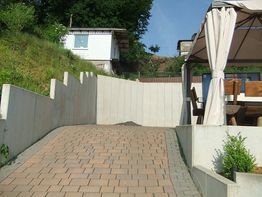 Auffahrt mit Pflastersteinen, links Böschung, rechts Pavillon mit Tisch und Stühlen, im Hintergrund Bäume.