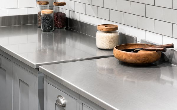 Encimera de cocina de acero inoxidable con acabado liso y reflectante, acompañada de tarros de vidrio con alimentos y un bol de madera sobre muebles blancos y salpicadero de azulejos.