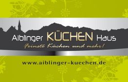 Grünes Schild des Aiblinger Küchenhauses mit Website und Bergsilhouette im Hintergrund.