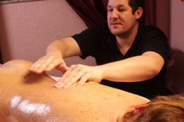 Person massiert den Rücken eines liegenden Menschen in einem Raum mit dunklen Vorhängen.