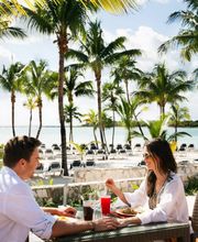 Pareja desayunando al aire libre con vista a la playa, rodeados de palmeras.