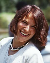 Eine lächelnde Frau mit braunen Haaren, trägt eine weiße Bluse und eine Halskette.