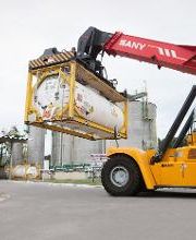 Guindaste amarelo movendo contêiner em área industrial com silos ao fundo.