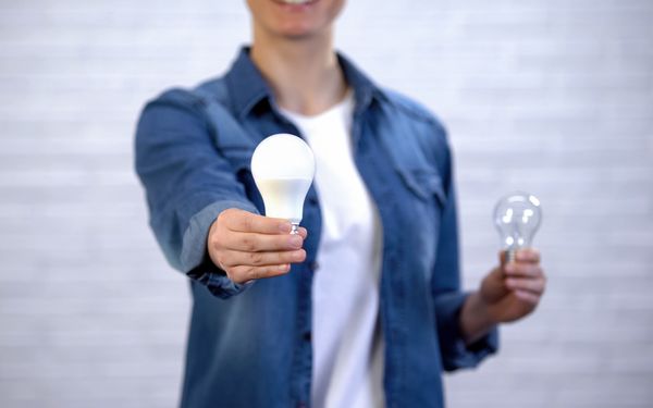 Eine Frau hält eine LED Lampe in die Kamera und eine Halogenlampe weiter hinten im Bild, um den tausch von Halogen auf LED zu symbolisieren.