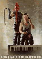 Ein Mensch in Latexkleidung hält eine Peitsche, ein anderer sitzt vor einem Keyboard mit Telefon.