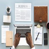 Typing on laptop with documents, coffee, notebook, and phone on a desk. Business newspaper visible.