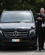 Ein Mann steht in Anzug neben einem schwarzen Mercedes-Großraumwagen auf einer Straße.