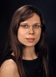 Frau mit langen braunen Haaren und Brille vor schwarzem Hintergrund, trägt ein schwarzes Oberteil.