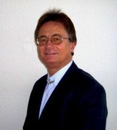 Ein Mann im Anzug mit Brille lächelt vor einer weißen Wand.