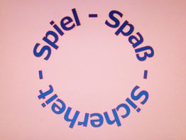 Runder Schriftzug mit den Wörtern "Spiel - Spaß - Sicherheit" auf rosa Hintergrund.
