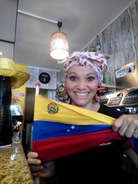 Mujer sonriente sostiene una bandera de Venezuela en un café, con decoración acogedora.