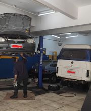 Carro em elevador mecânico com capô aberto; mecânico trabalha; Kombi no chão ao lado.