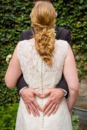 Braut im weißen Kleid von hinten, Rücken wird von Bräutigam umarmt. Sie formen ein Herz.