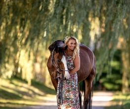 Frau in Blumenkleid steht mit Pferd auf einem von Bäumen gesäumten Weg.