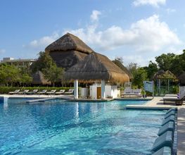 Piscina amplia con tumbonas y edificios de techo de paja rodeados de vegetación.