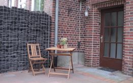 Holzmöbel auf Terrasse vor Backsteinwand, Tisch mit zwei Pflanzen, neben Gitter mit Steinen.