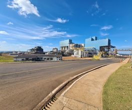 Fábrica industrial em dia ensolarado, céu azul com algumas nuvens, cercada por estrada asfaltada.