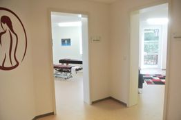 Flur mit zwei offenen Türen, rote Grafik an der Wand, Raum mit Massageliege und Bild sichtbar.