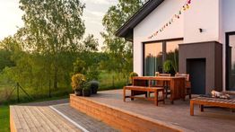 Modern patio with wooden deck, furniture, and string lights, surrounded by lush greenery.
