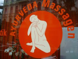 Schaufenster mit orangefarbener Grafik für Ayurveda-Massagen, umgeben von Pflanzen.