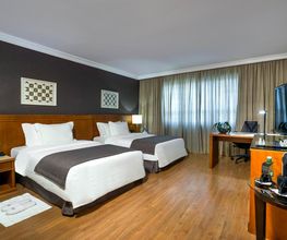 Quarto de hotel com duas camas, mesa, TV e janelas com cortinas num ambiente aconchegante.