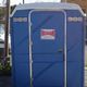 Bagno chimico blu all'aperto con un cartello di noleggio. Furgone bianco parcheggiato a sinistra.