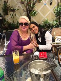 Duas mulheres sorrindo em mesa de café ao ar livre, com copo de suco e narguilé à frente.