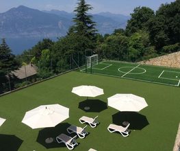 Liegestühle und Sonnenschirme auf einem Platz mit Kunstrasen, dahinter ein kleiner Fußballplatz.
