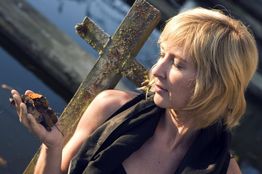 Vrouw met kort blond haar kijkt naar een roestige kruisbeeld bij een rivier. Ze houdt bladeren vast.