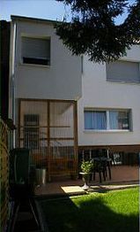 Zweistöckiges Gebäude mit Terrasse und Garten, Stühle und Tisch im Schatten von Bäumen.
