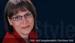 Eine Frau mit braunem Haar und Brille, trägt ein rotes Oberteil, lächelt vor dunklem Hintergrund.