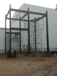 Estructura de acero en construcción junto a un edificio blanco con andamios alrededor.
