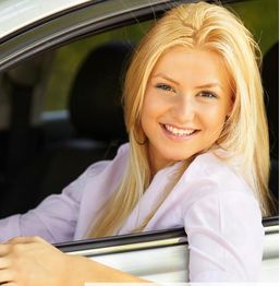 Smiling woman with blonde hair in a car, looking out of the window on a sunny day.