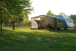 Wohnwagen im Grünen mit zwei Menschen unter Vorzelt, umgeben von Bäumen und Sonnenschein.
