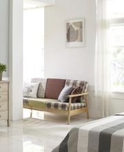 Habitación luminosa con cama, sofá a cuadros, cómoda con planta y ventana grande.