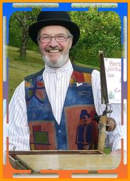 Ein lächelnder Mann mit Hut und bunter Weste hält ein Musikinstrument im Freien.