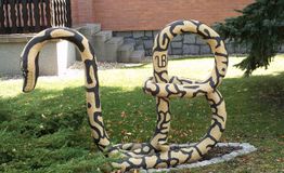 Eine große, gelbe Skulptur einer Schlange in "B"-Form auf einem Rasen vor einem Haus.