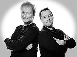 Zwei Männer lächeln, mit verschränkten Armen, in schwarz-weißem Foto vor hellgrauem Hintergrund.