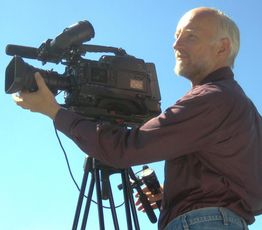 Älterer Mann mit Bart filmt mit großer Videokamera bei klarem Himmel.